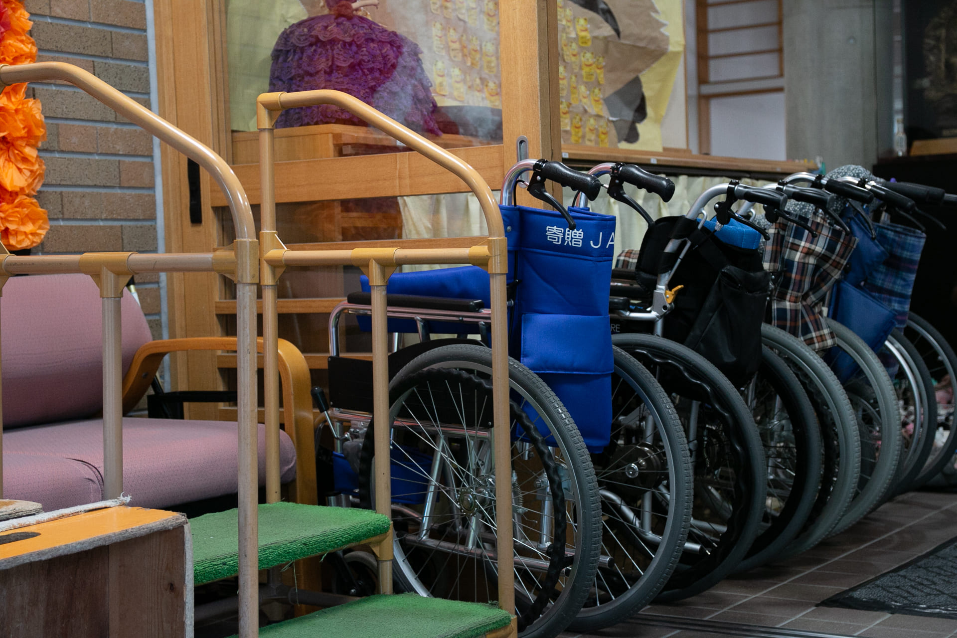 介護施設のロビーに整列した福祉用車いすと手すり付き椅子が配置されている。
